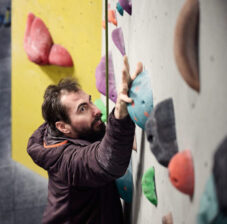 A man reaches for a hold on a climbing wall. He is wearing a burgundy hoodie and has dark hair, a beard and a moustache.