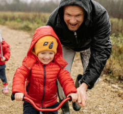 Image shows a smiling family on a walk in the countryside. In the foreground a father pushes a child of a bicycle and in the background a mother holds the hand of a younger child.