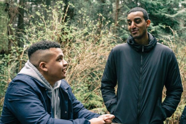 Two men are outdoors in a green space speaking to one another.