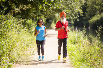 Two people run down a path in the sunshine, surrounded by greenery.