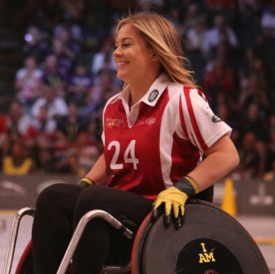 A smiling woman with blond hair sits in her wheelchair on a basketball court. She is wearing gloves and a number 24 team shirt.