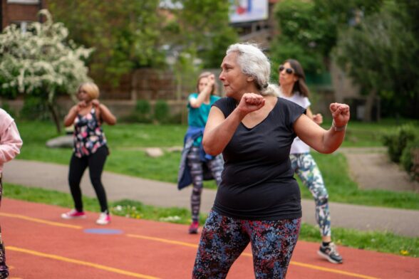 An older woman participates in an outdoor exercise class at a running track. Other women are exercising in the background.