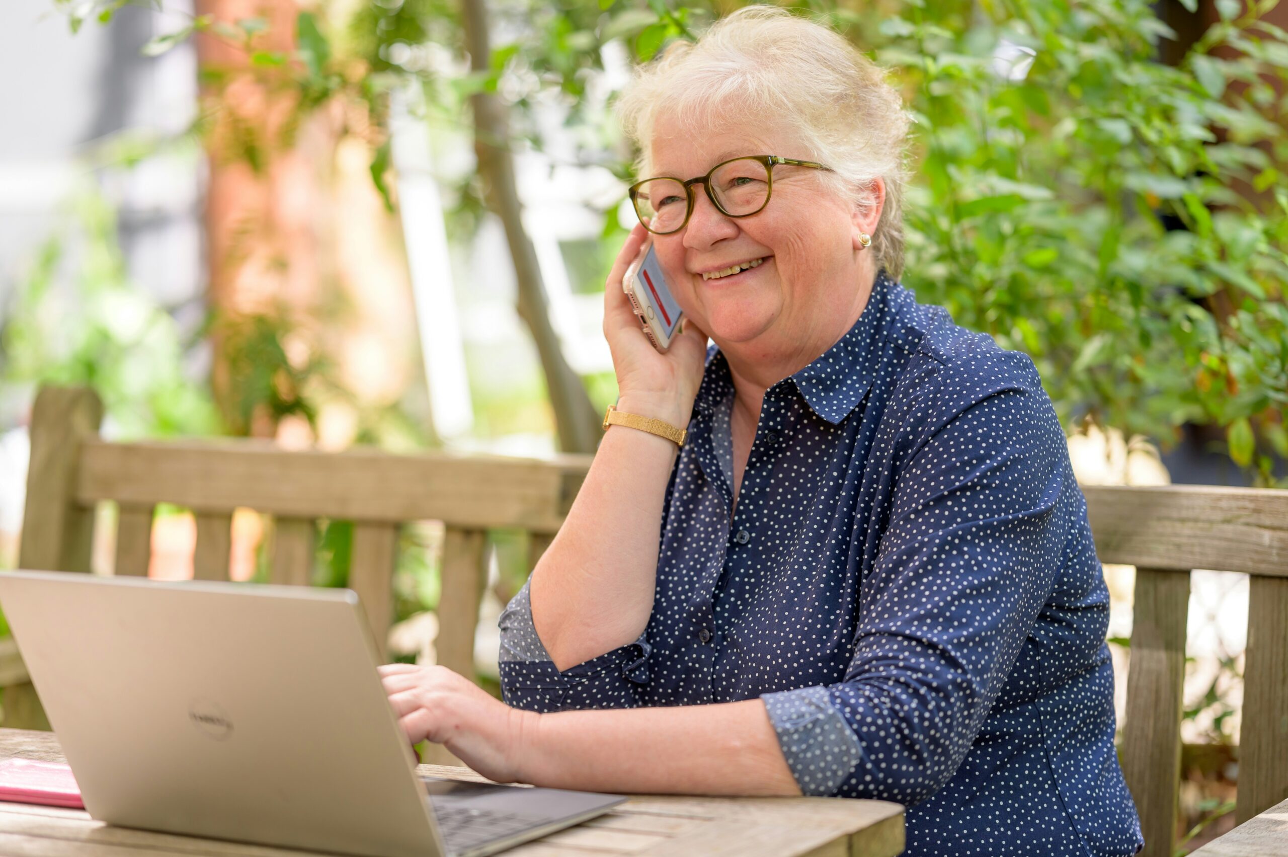 A woman sits at an outdoor table with greenery behind her. Her laptop is open in front of her and she smiles while holding a phone to her ear.