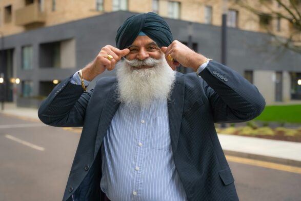 A man does a double thumbs up to to camera. He is smiling and wearing a dark suit over a lightblu shirt. He has a large white beard, bushy moustache and is wearing a turban.