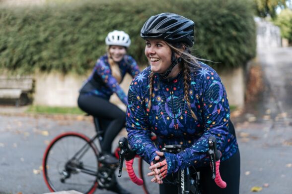 Two women on bicycles are smiling. They are wearing helmets and cycling around a paved area surrounded by greenery.