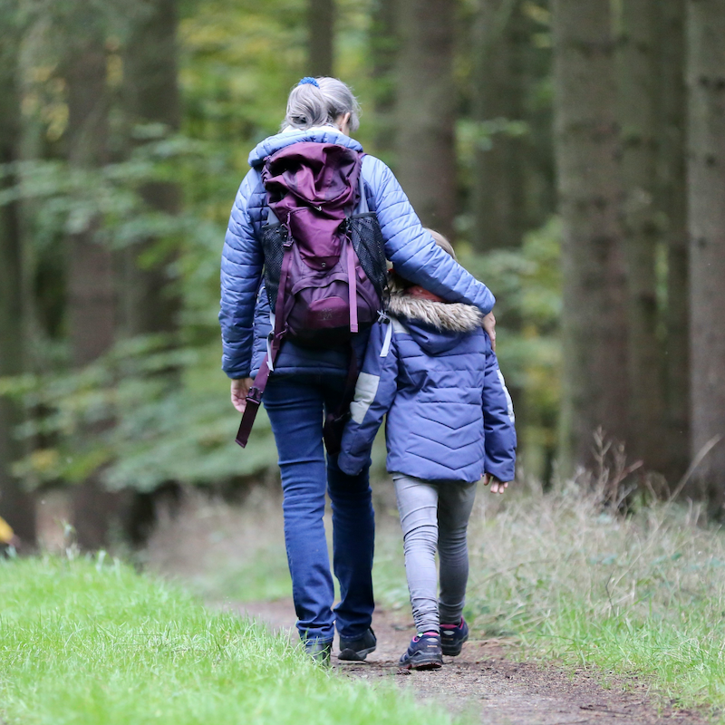 A woman and a child walk along a path in a forest with their backs to the camera. Both are wearing puffer jackets and walking boots. The woman has a hiking backpack on and has very arm around the shoulders of the child.
