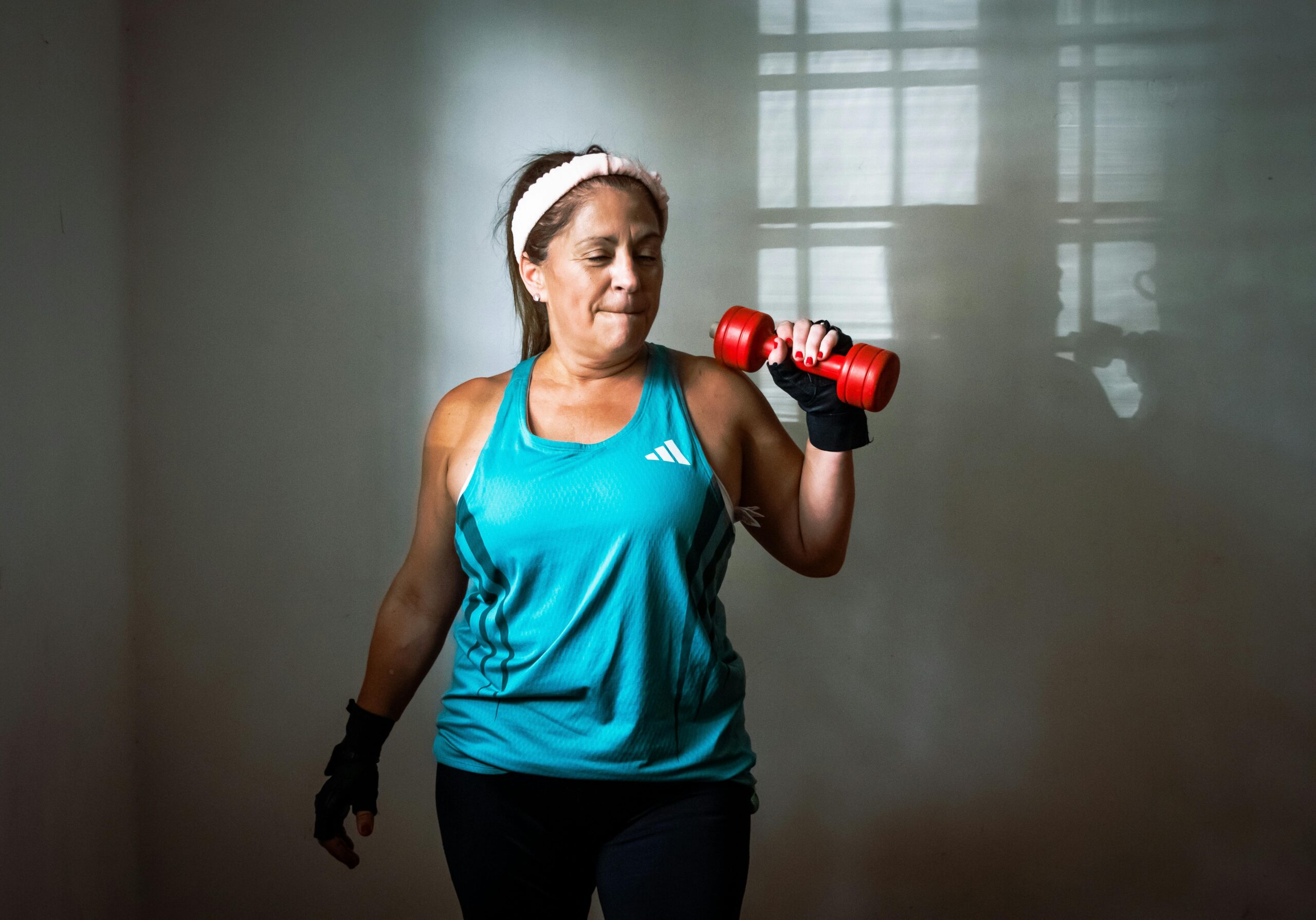 A woman smiles while lifting a weight in her right hand. She is wearing a white sports headband, a turquoise tank top and black leggings.