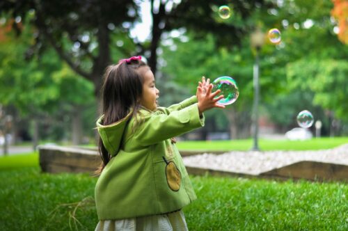 A little girl with dark hair is wearing a green rain jacket. She is standing in a lush green park and reaching out for a large bubble floating in the air.