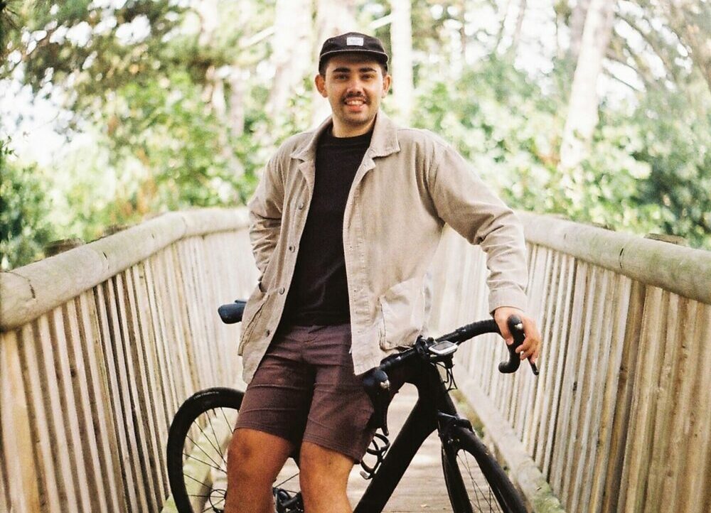 A man leans on his bicycle while standing on a wooden bridge. he is wearing a baseball cap and wearing a beige jacket over a black t-shirt and grey shorts.