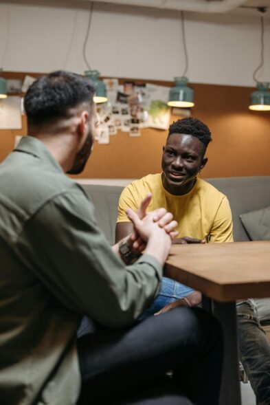 Two men sit at a table in a cafe-style space. They are leaning towards each other, listening carefully and one man is gesticulating.