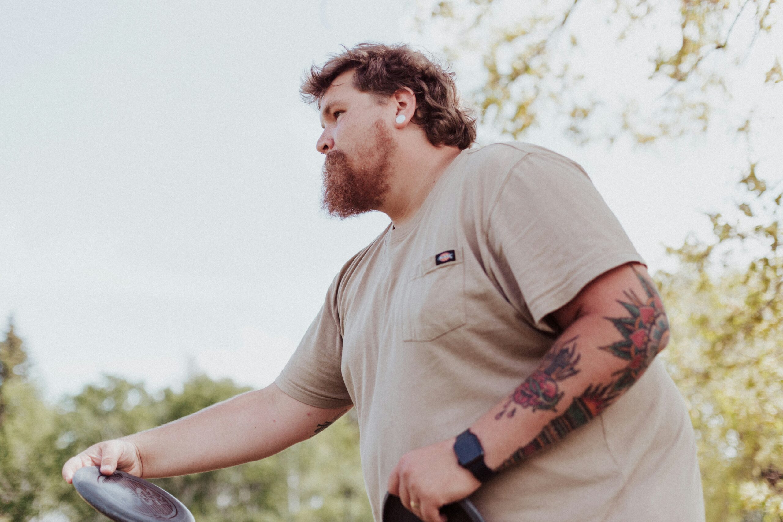 A man with a short beard positions himself to throw a discus. He has a tattooed left arm and is wearing a beige t-shirt. He holds the discus in his right hand.