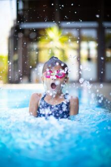 A child surfaces in a swimming pool, causing water to splash upwards. She is wearing pink goggles a blue swimsuit and is making a surprised expression.