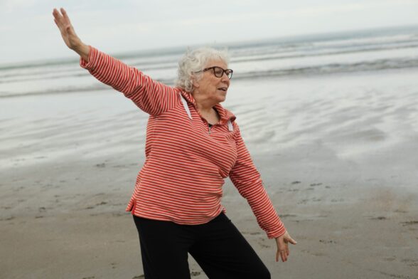 An older woman walks on the sand at the edge of the sea. She spreads her arms as though she is flying. She wears a red and white stripy long sleeve top and black trousers.